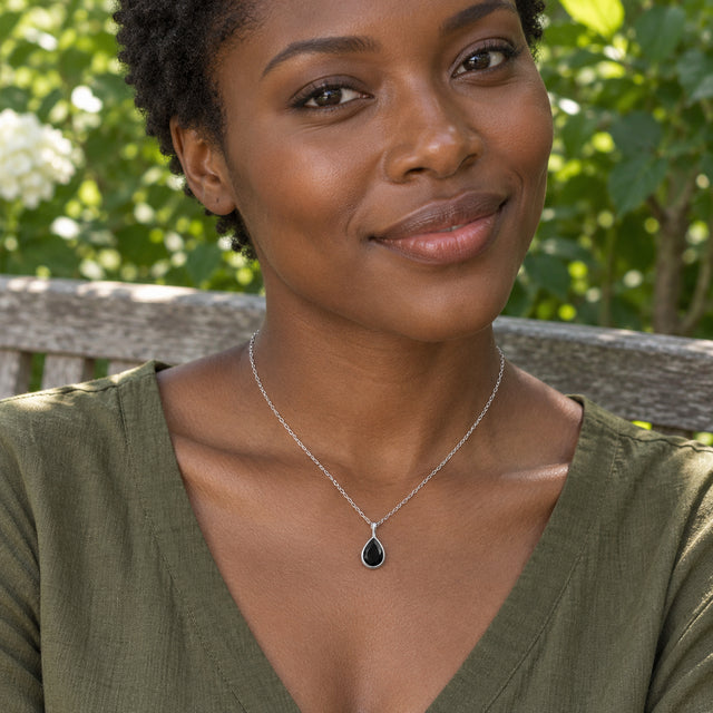 A woman in an olive V-neck top wears a silver black-stone pendant while seated on a wooden garden bench.