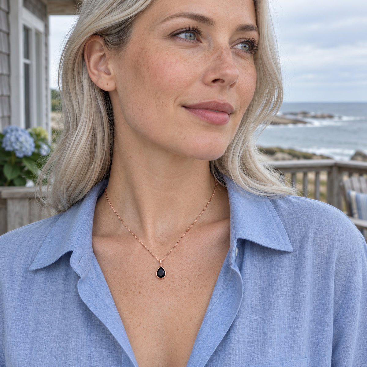 A blonde woman in a blue shirt wears a rose gold black pear pendant on a veranda overlooking the sea.