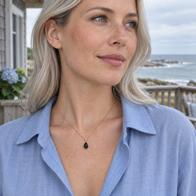 A blonde woman in a blue shirt wears a rose gold black pear pendant on a veranda overlooking the sea.