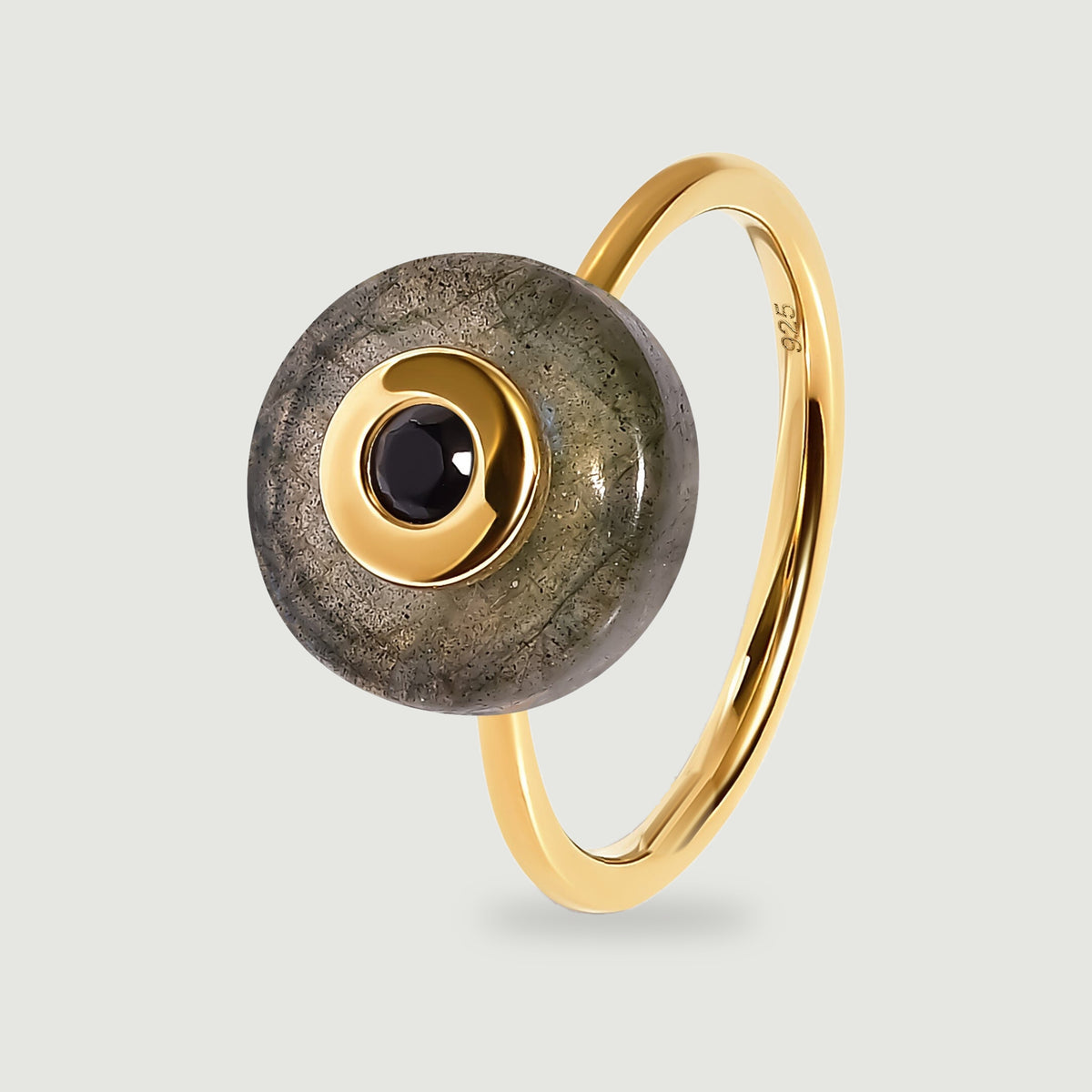 A yellow gold vermeil ring holds a round grey labradorite disc with a black central stone, shown upright on a light grey surface.