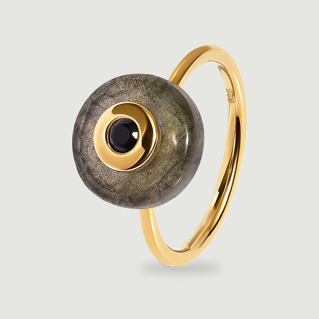 A yellow gold vermeil ring holds a round grey labradorite disc with a black central stone, shown upright on a light grey surface.