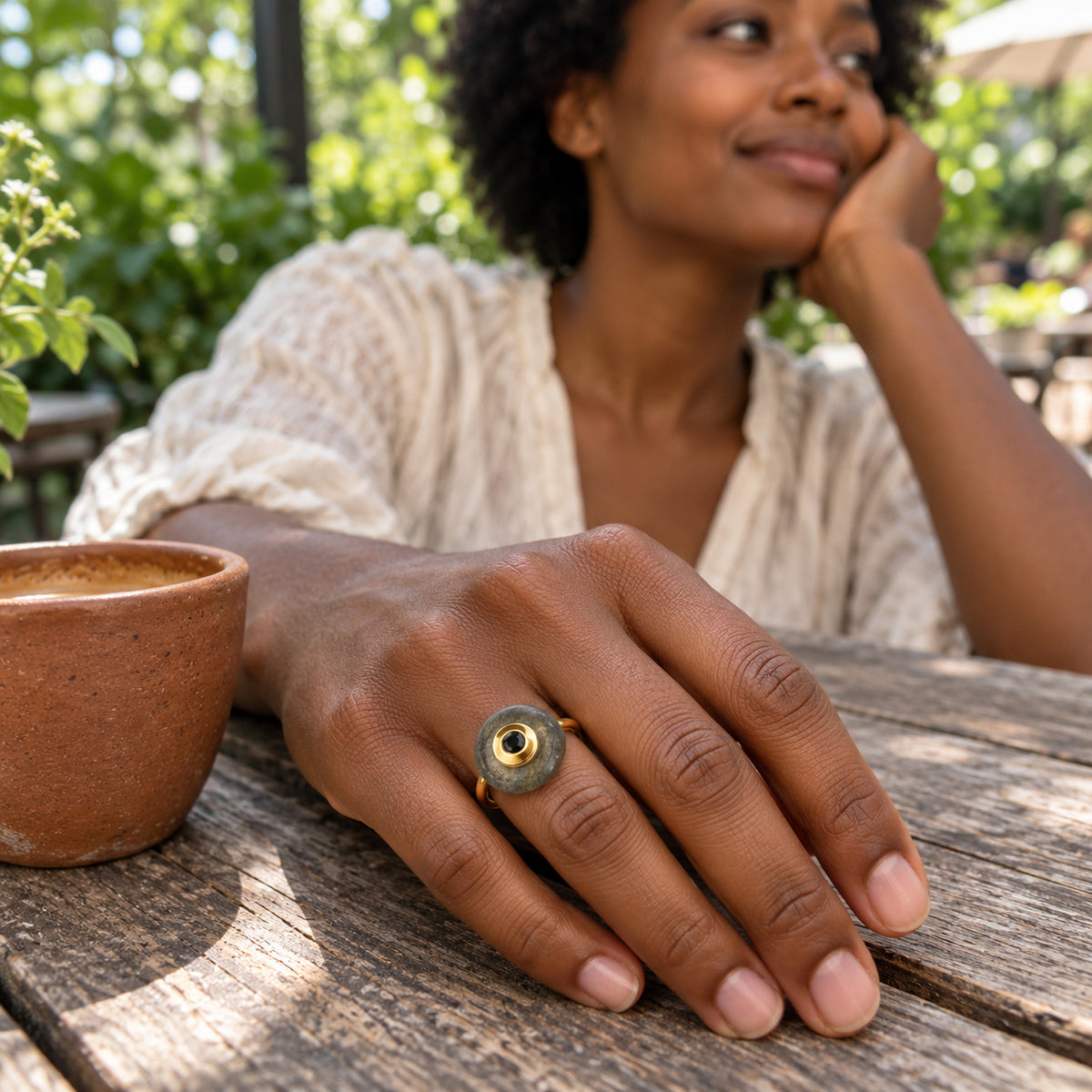 A woman wears the yellow gold vermeil labradorite ring on her hand beside a clay cup on a wooden cafe table.