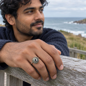 A man wears the sterling silver labradorite ring while resting his hand on a weathered wooden railing above the sea.