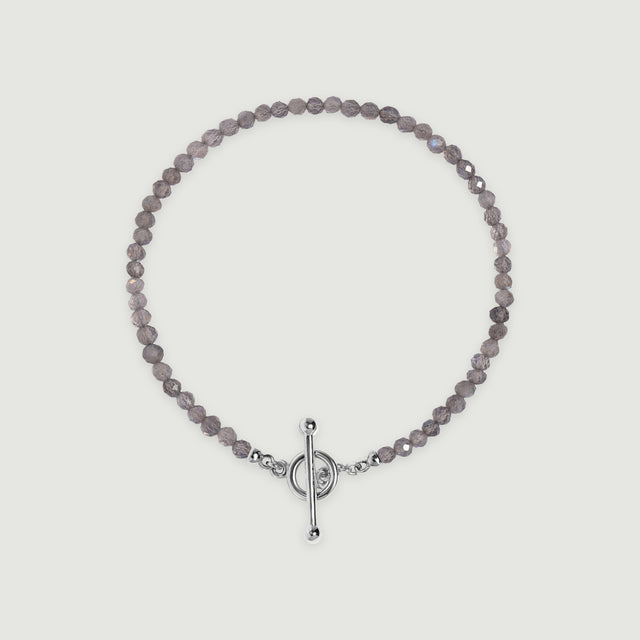 A complete grey labradorite bead bracelet with a polished silver ring and bar clasp lies centered on a light grey surface.