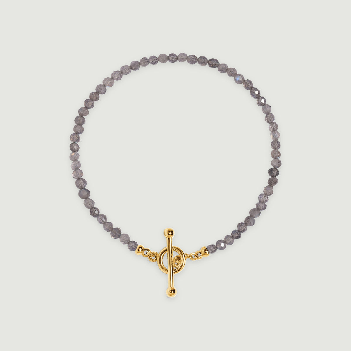 Faceted grey labradorite beads form an oval bracelet with a yellow gold ring and bar clasp on a light grey surface.