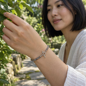 A woman with a black bob wears the grey bead bracelet with a silver ring and bar clasp while touching a green leaf in a garden.