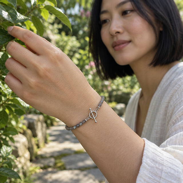 A woman with a black bob wears the grey bead bracelet with a silver ring and bar clasp while touching a green leaf in a garden.