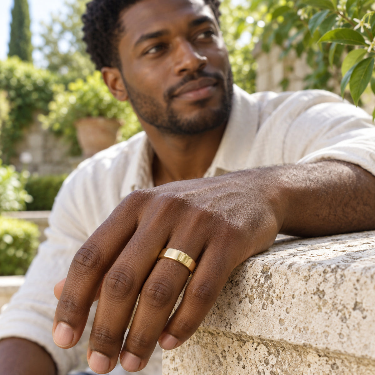 A yellow gold band is worn on a man's hand resting on a stone ledge in a leafy courtyard.
