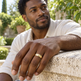 A yellow gold band is worn on a man's hand resting on a stone ledge in a leafy courtyard.