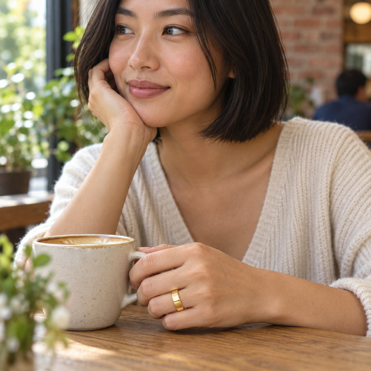A woman wears a yellow gold band while holding a ceramic coffee cup at a wooden cafe table.