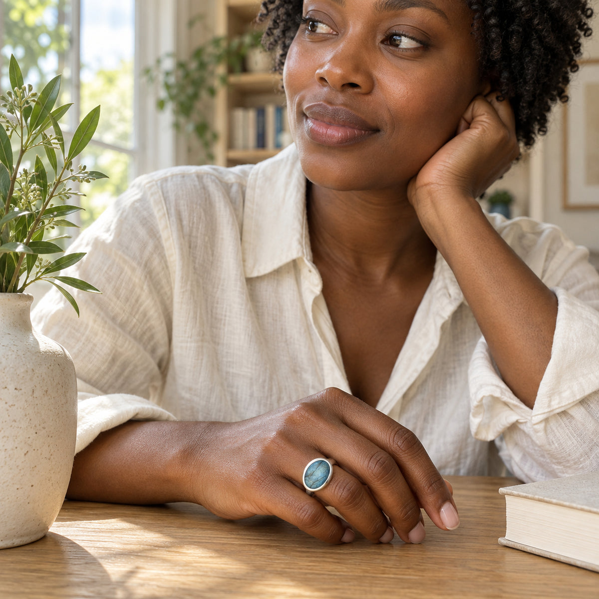 A silver ring with an oval blue labradorite is worn on a hand resting on a wooden table beside a ceramic vase.