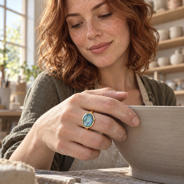 A yellow gold vermeil ring with a veined blue stone is shown on a hand holding the rim of a pottery bowl.