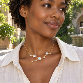 A woman with short curls wears a yellow gold moonstone necklace above a white linen shirt in a leafy stone courtyard.