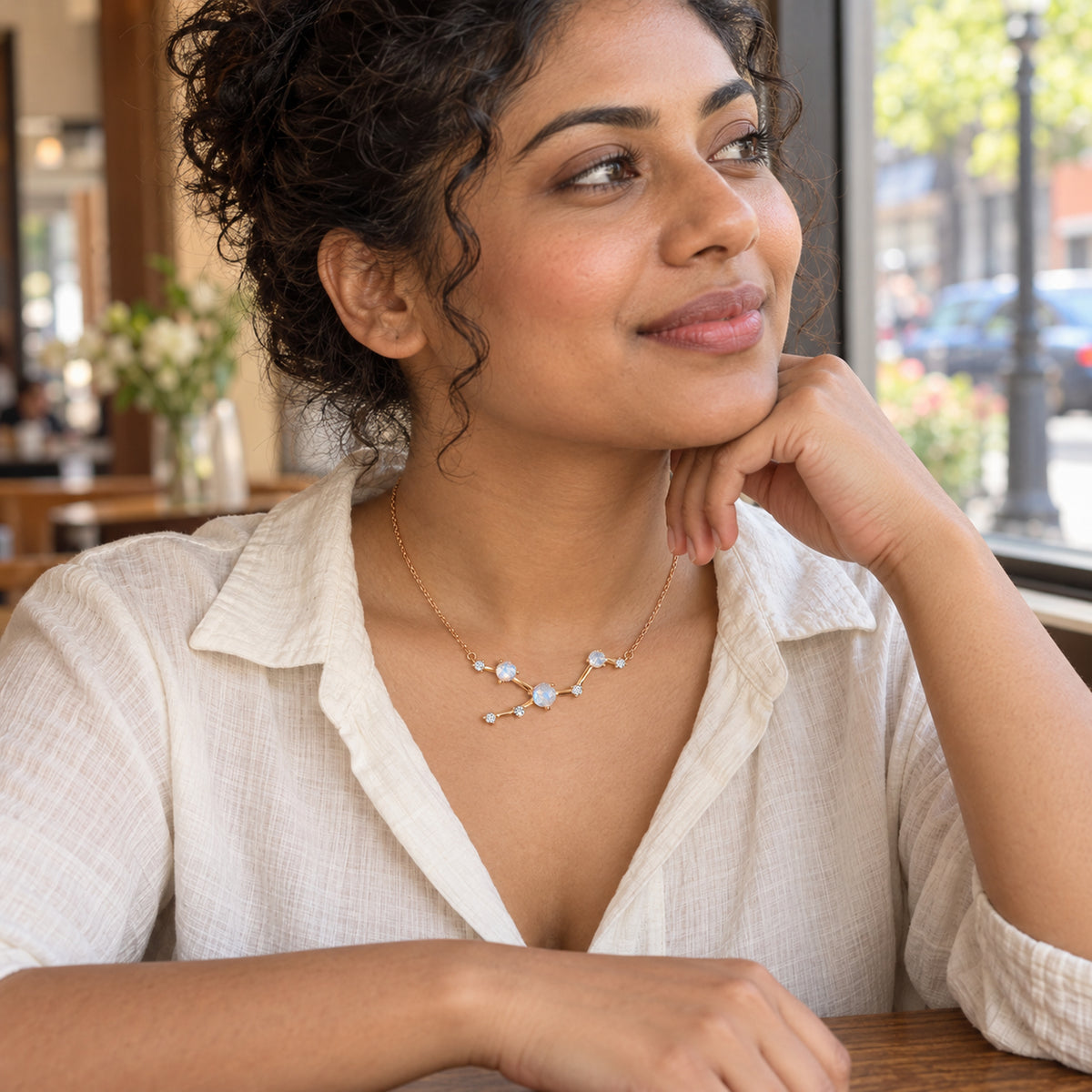 A woman rests her chin on her hand while wearing a rose gold moonstone necklace beside a cafe window.