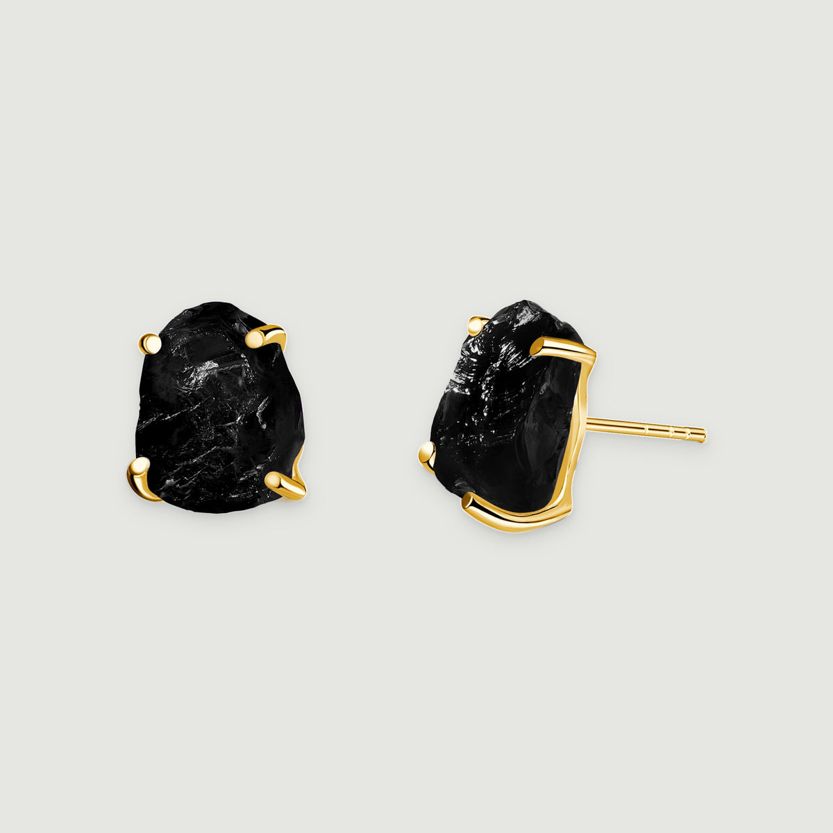 Two raw black obsidian stud earrings with yellow gold prongs rest on a light gray surface.