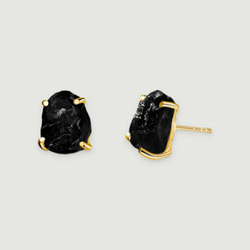 Two raw black obsidian stud earrings with yellow gold prongs rest on a light gray surface.