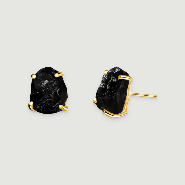 Two raw black obsidian stud earrings with yellow gold prongs rest on a light gray surface.