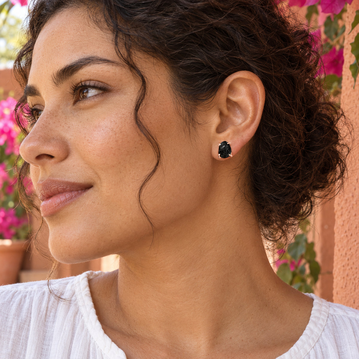 A woman with curly dark hair wears a rose gold black obsidian stud beside a terracotta wall and pink flowers.