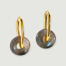 A pair of polished yellow gold hoops carries round gray labradorite discs on a light gray surface.