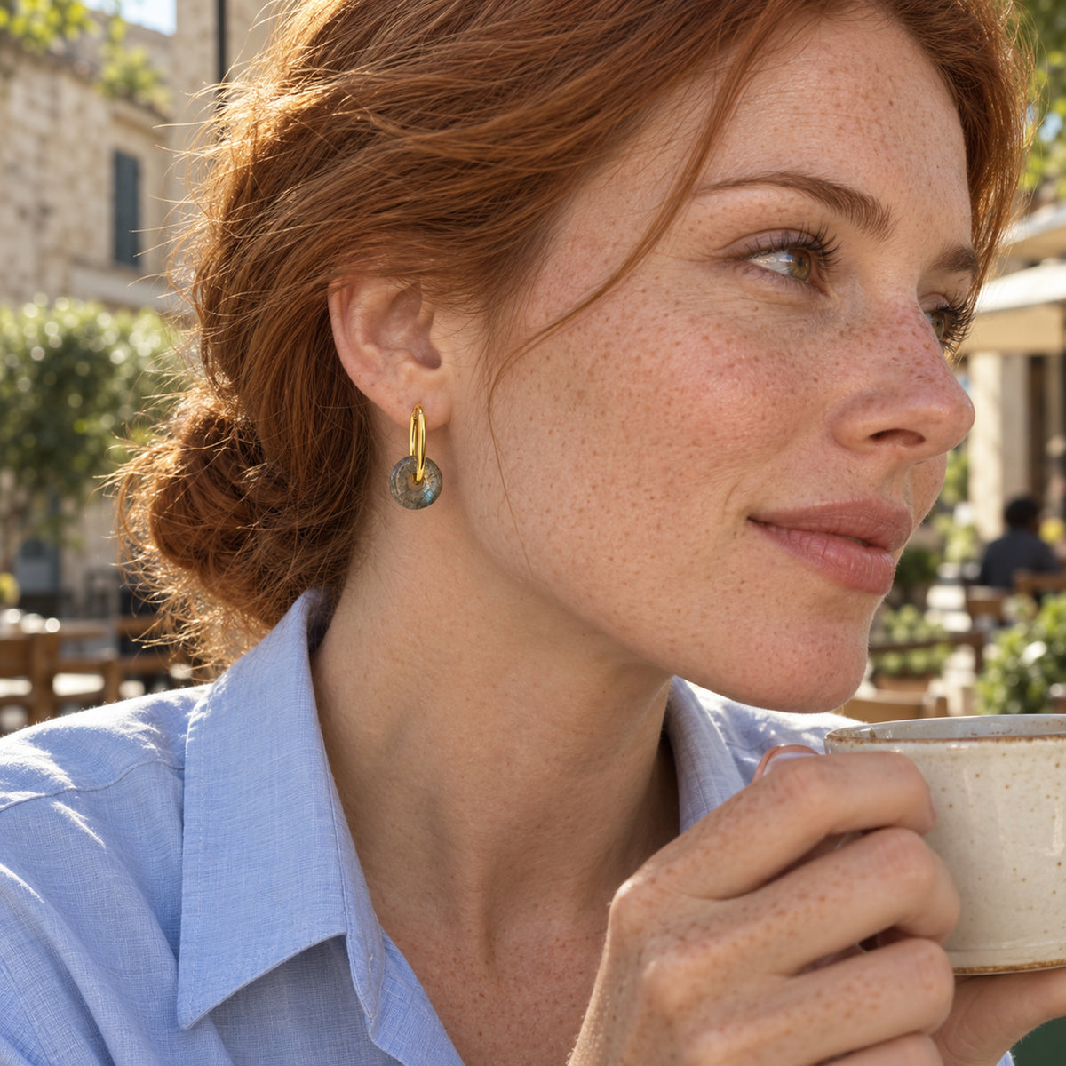 A freckled woman with red hair wears a yellow gold labradorite hoop while holding a ceramic cup at an outdoor cafe.