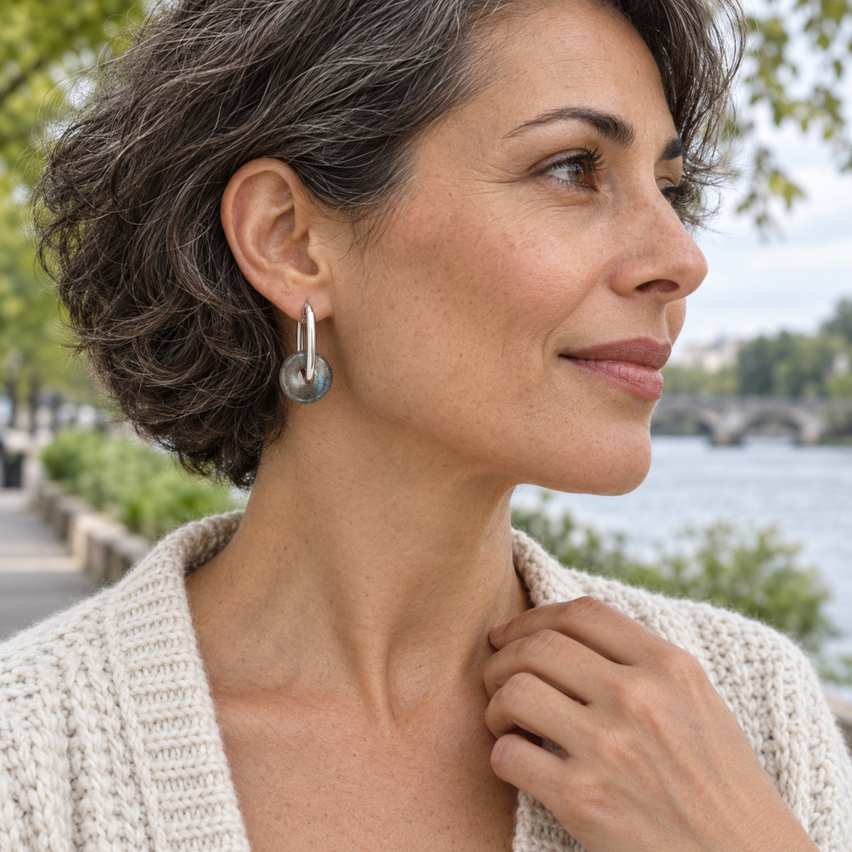 A woman with short salt-and-pepper hair wears a silver-tone labradorite hoop beside a river.
