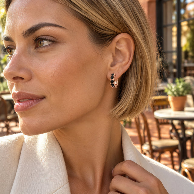 A woman with a blonde bob wears a rose gold black onyx hoop earring at a sunlit cafe terrace.