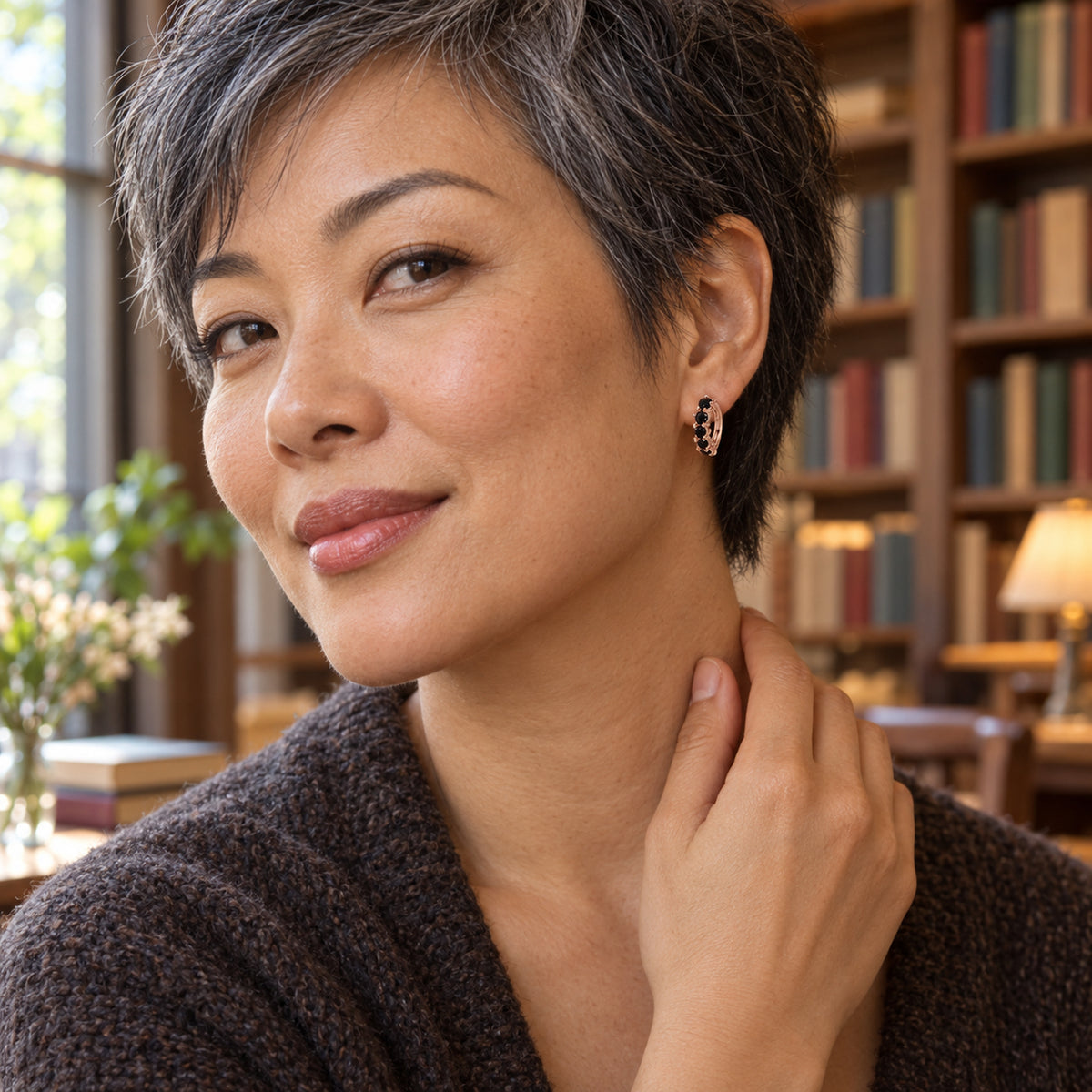 A woman with short salt and pepper hair wears a rose gold black onyx hoop earring beside bookshelves.