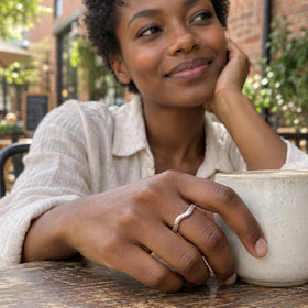 A silver textured ring is worn on a hand beside a ceramic cup in a brick cafe courtyard.