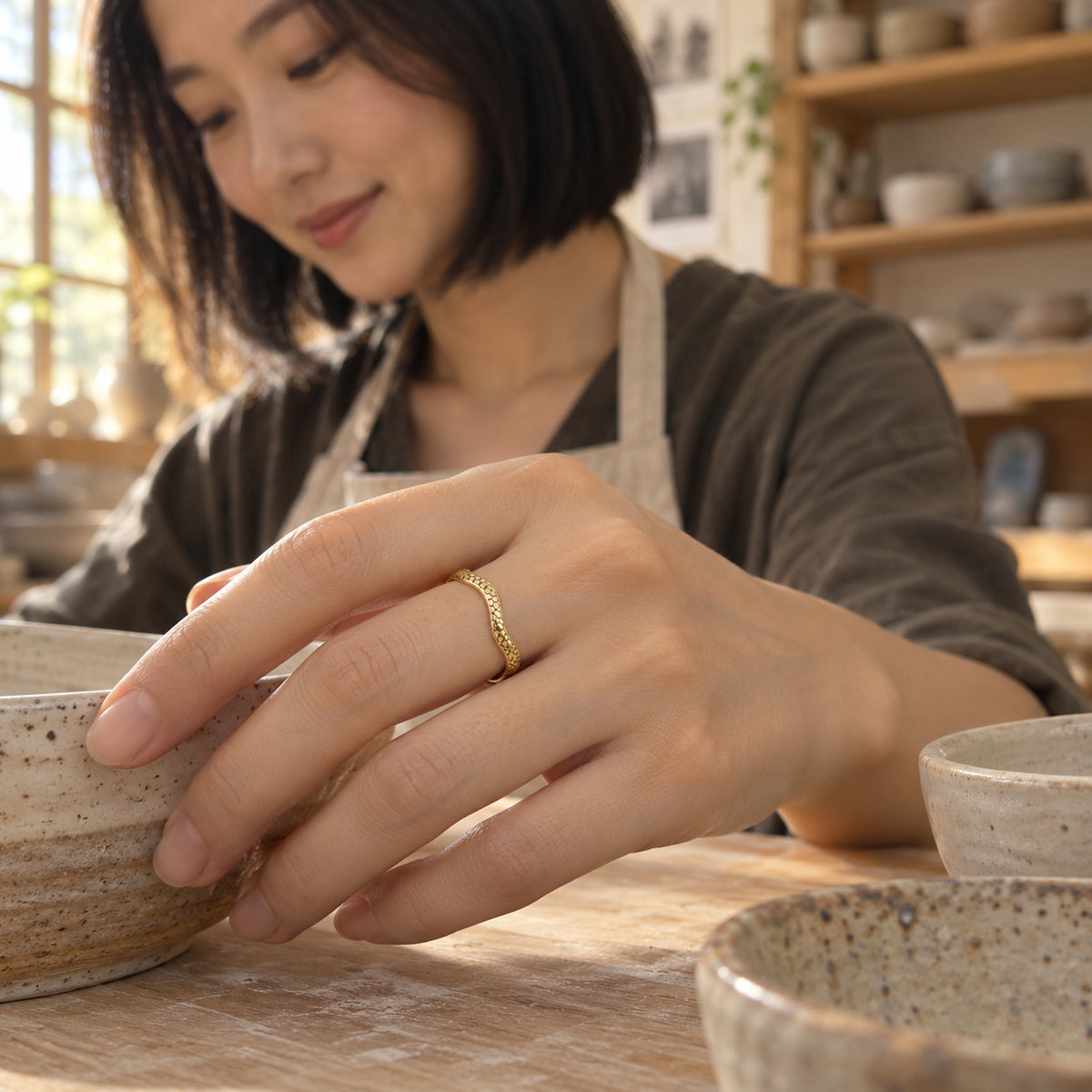 A yellow gold textured ring is worn on a hand beside ceramic bowls in a pottery studio.