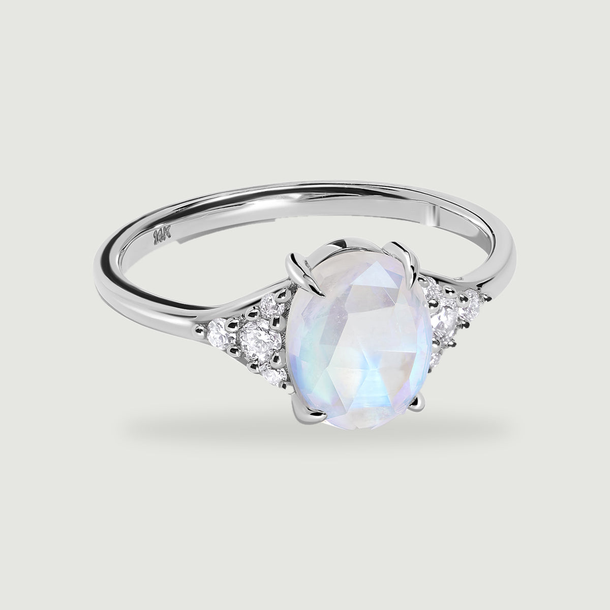 An oval faceted moonstone and clustered clear diamonds sit in a polished white gold ring on a pale gray surface.