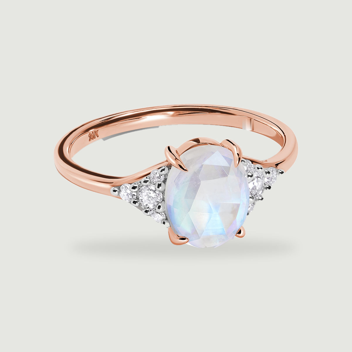 A rose gold ring with an oval moonstone and diamond shoulders casts a soft shadow on a light gray background.