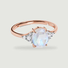 A rose gold ring with an oval moonstone and diamond shoulders casts a soft shadow on a light gray background.