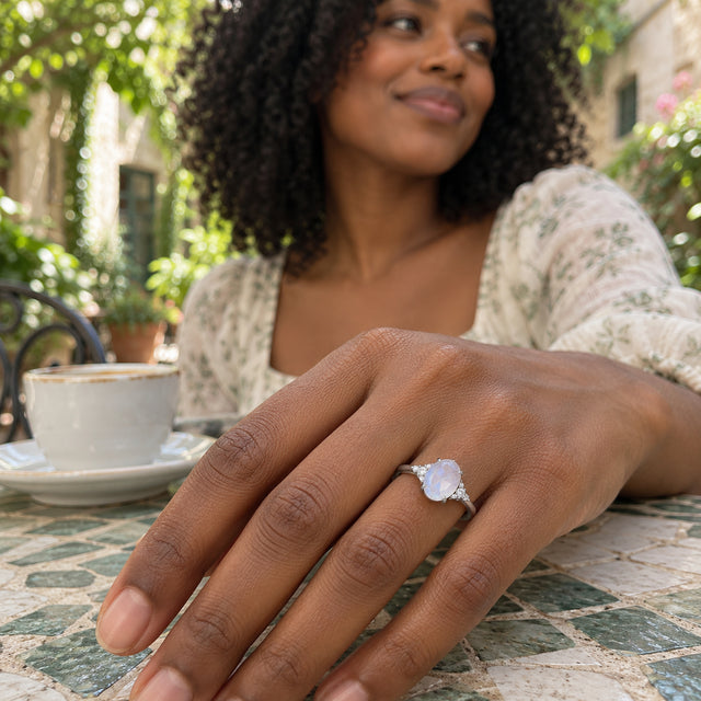 A white gold moonstone and diamond ring is worn on a hand resting beside a coffee cup on a tiled courtyard table.