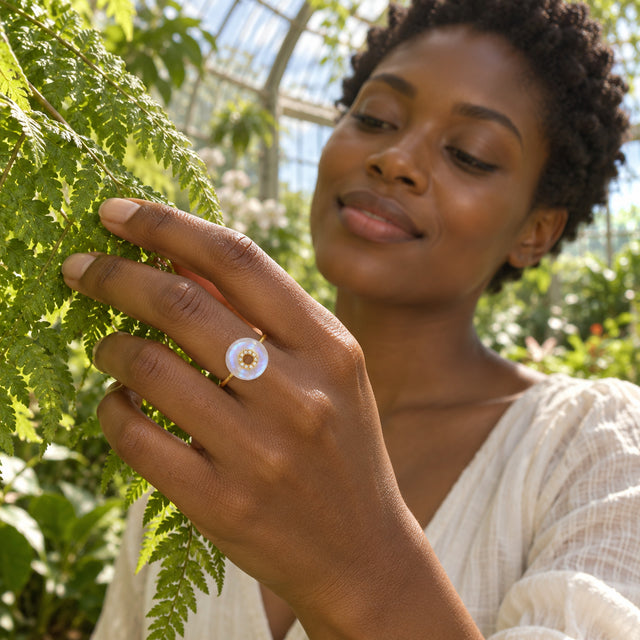 A yellow gold moonstone ring is worn on a hand touching fern fronds in a sunlit conservatory.