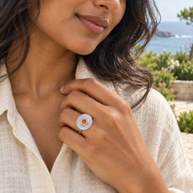 A rose gold moonstone ring is shown on a raised hand beside a cream linen shirt with sea and greenery behind it.