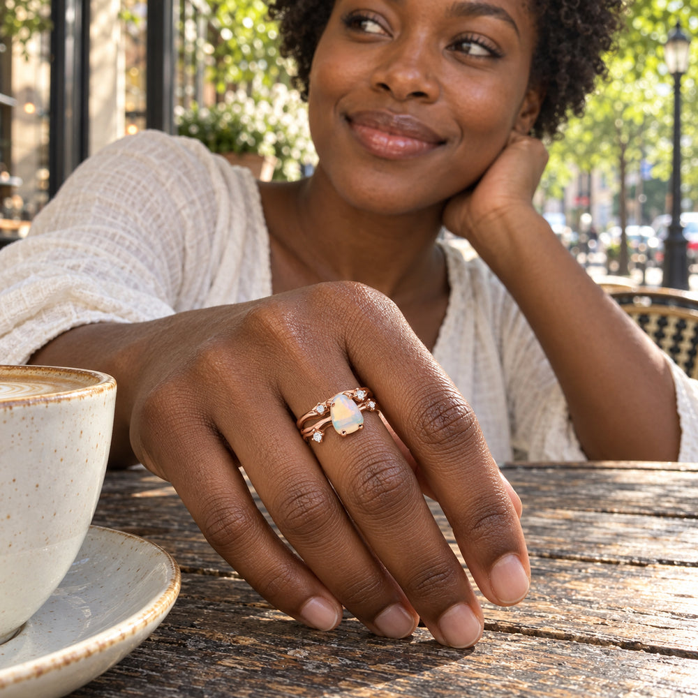 A rose gold opal ring is worn on a hand resting on a weathered cafe table beside a ceramic cup.