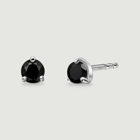Two round faceted black obsidian studs show polished silver prongs and a straight earring post on a light gray surface.