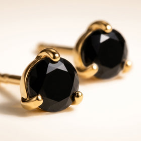 The polished yellow gold setting and angular black stone facets fill a close view of two stud earrings on a cream surface.