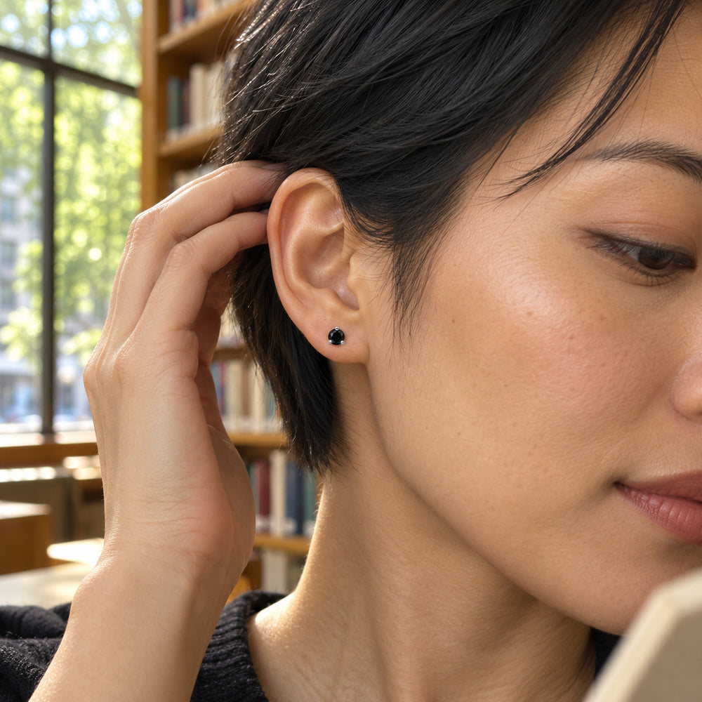 A tiny black obsidian stud with silver prongs is worn beside short dark hair, with sunlit windows and bookshelves behind it.