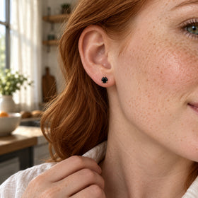 A round black obsidian stud in rose gold prongs sits on a freckled ear beside red hair in a bright kitchen.