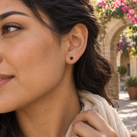 A black obsidian stud with yellow gold prongs is worn beside dark wavy hair against a stone arch and flowering vines.