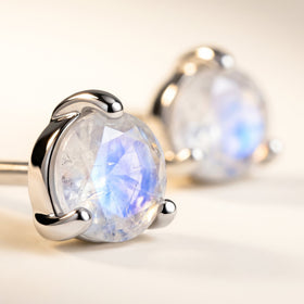 A silver three-prong stud holds a faceted moonstone with blue reflections, with a second earring softly blurred behind it.