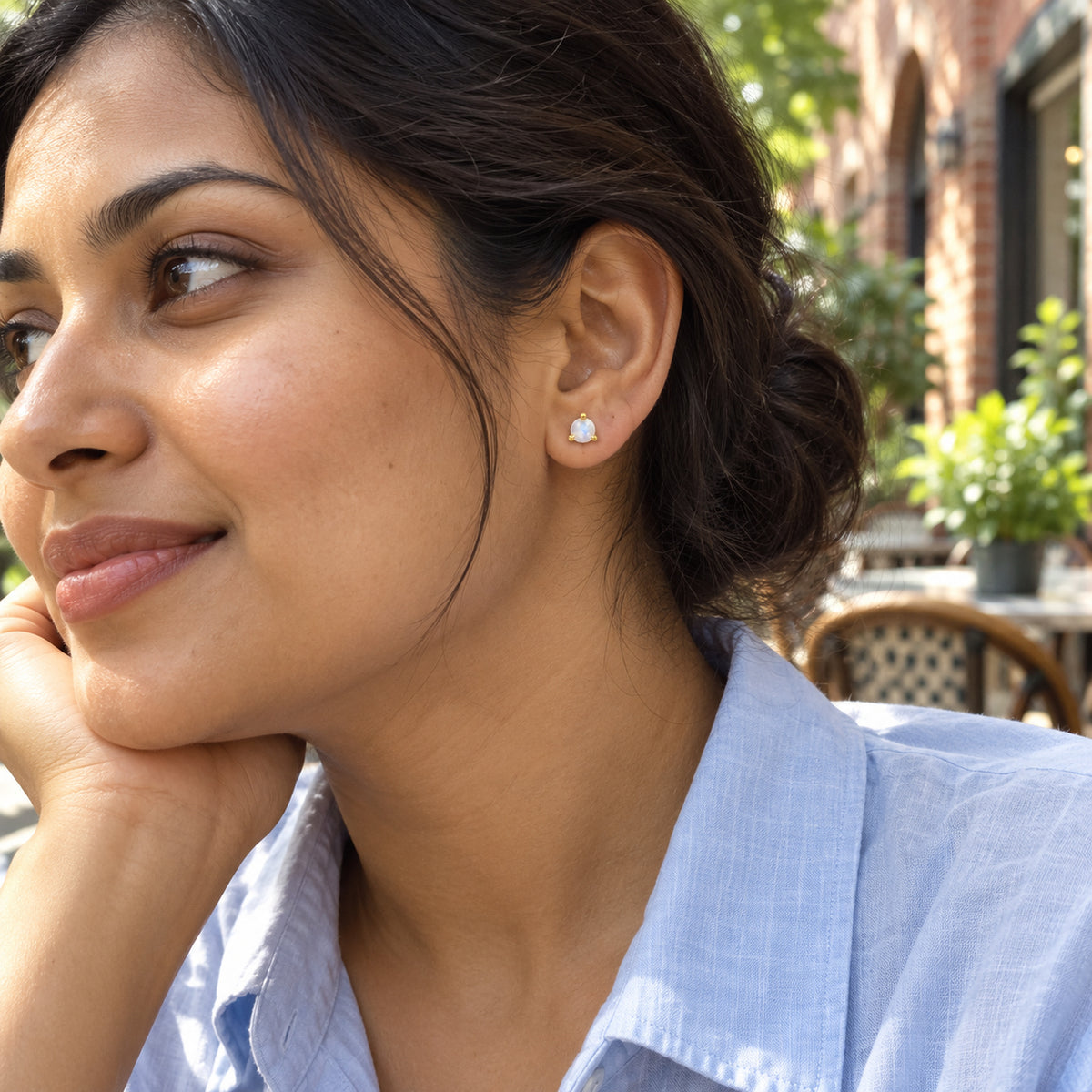 A small yellow gold vermeil moonstone stud sits on a model's earlobe at an outdoor cafe, above a pale blue shirt.
