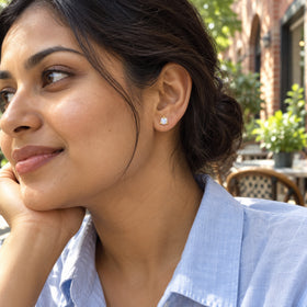 A small yellow gold vermeil moonstone stud sits on a model's earlobe at an outdoor cafe, above a pale blue shirt.