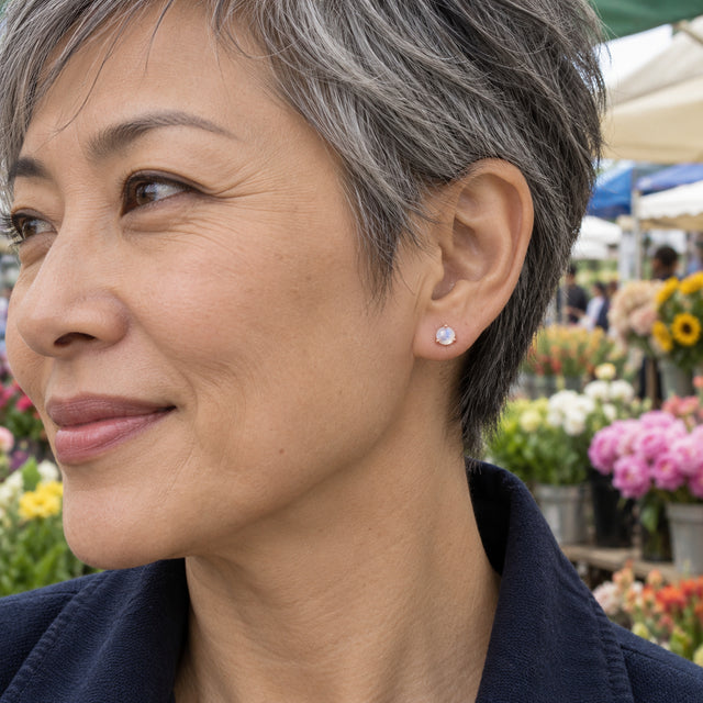A rose gold vermeil moonstone earring is shown beside short gray hair, with flower stalls softly blurred behind the model.