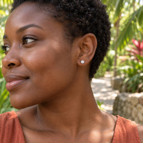 Warm yellow gold prongs hold a pale moonstone on an earlobe above a terracotta top, against leafy garden scenery.
