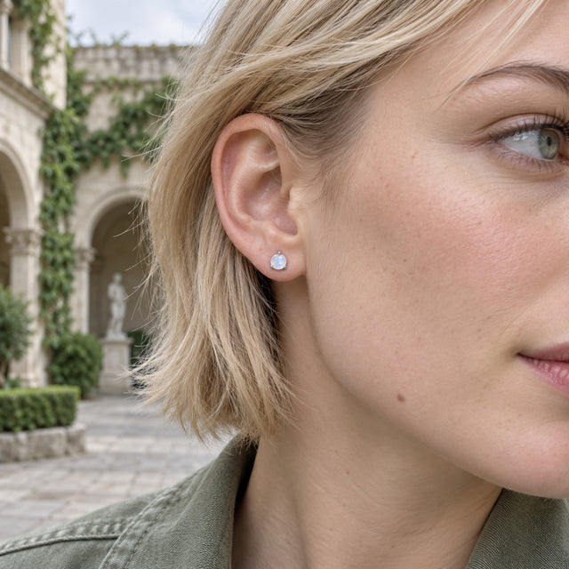 A white gold moonstone stud is shown on an exposed ear beside blond hair, with stone arches and vines in the courtyard background.