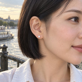 A blush rose gold moonstone stud rests on an earlobe beside a black bob, with a river and boats behind the model in evening light.