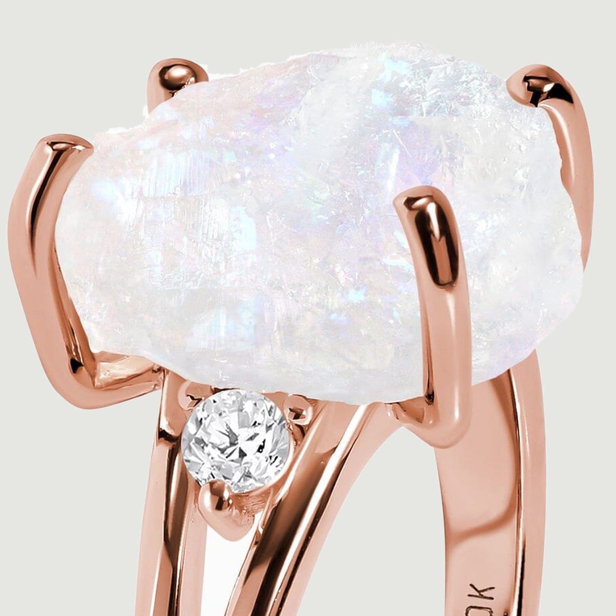 The raised rose gold setting surrounds a rough moonstone above a small round clear side stone.