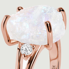 The raised rose gold setting surrounds a rough moonstone above a small round clear side stone.